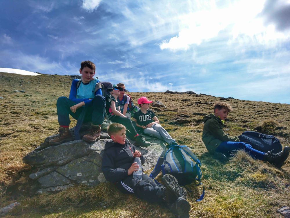 A group of people sit and rest on a grassy hillside during a hike. Some are sitting on a large rock while others sit on the ground beside their backpacks. The sky above is bright with scattered clouds, and patches of snow are visible on the higher slope in the background.
